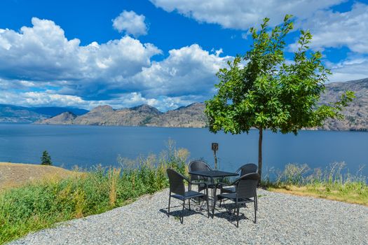 Dining table and chairs on picnic area with panoramic view at Okanagan lake and lonely tree by side