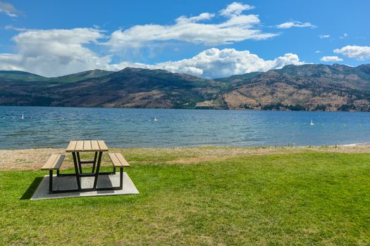 Picnic table with benches on Okanagan lake shore. Table and benches at rest area close to waterfront with mountains view on cloudy and blue sky background.