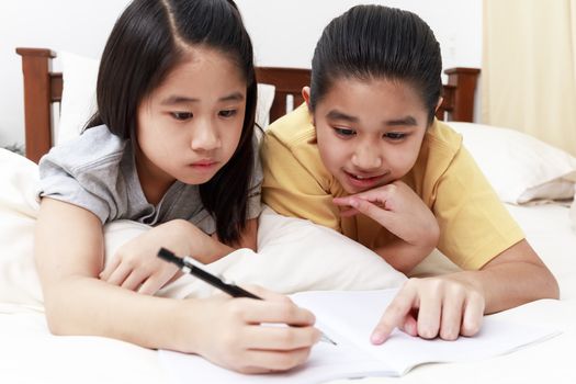Asian little girls doing homework together on bed in bedroom. Asian students learning at home. Asian elder sister teaching little sister to doing homework.