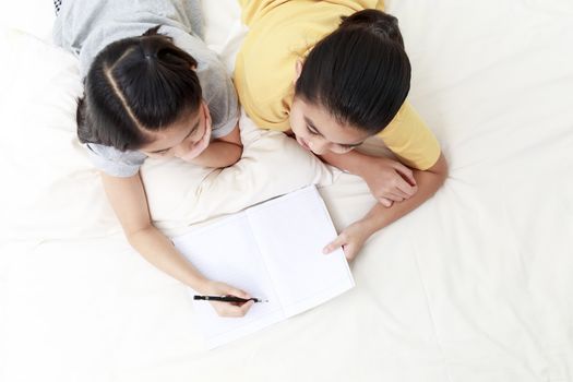 Top view Asian little girls doing homework together on bed in bedroom. Asian students learning at home. Asian elder sister teaching little sister to doing homework.