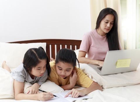 Asian family mother use laptop online working and her daughters do homework on bed in bedroom. Business women woking with laptop at home with her two daughters do home work together.