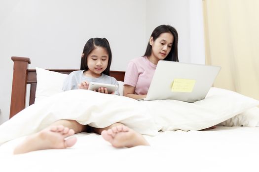 Asian mother and daugther looking laptop and tablet on bed in bedroom. Business women woking with laptop at home with her daugther smiling enjoy with tablet on bed. Work at home concept.