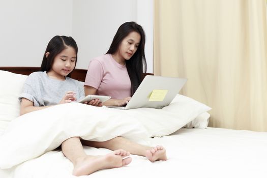 Asian mother and daugther looking laptop and tablet on bed in bedroom. Business women woking with laptop at home with her daugther smiling enjoy with tablet on bed. Work at home concept.