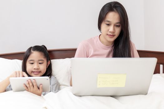 Asian mother and daugther looking laptop and tablet on bed in bedroom. Business women woking with laptop at home with her daugther smiling enjoy with tablet on bed. Work at home concept.