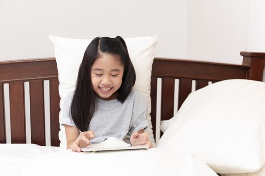 Asian little girl smiling in hands holding and play tablet on bed in bedroom. Little girl enjoy with tablet on bed. Asian girl looking and play tablet.