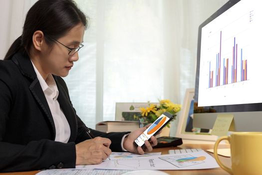 Business women holding smart phone check pie chart and write notes on desk at office. Business people analysis chart  report on phone and computer pc at home.