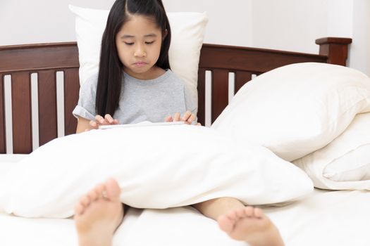 Asian little girl smiling in hands holding and play tablet on bed in bedroom. Little girl enjoy with tablet on bed. Asian girl looking and play tablet.