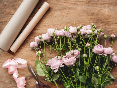 Top view on wooden table with roses, scissors, craft paper and pink ribbon on it. Florist work place. Accessories for making bouquets and floral compositions.