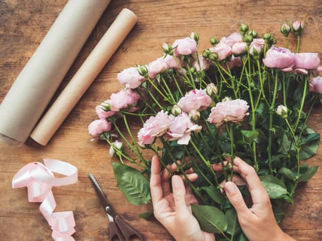 Top view on wooden table with roses, scissors, craft paper and pink ribbon on it. Florist work place. Accessories for making bouquets and floral compositions.