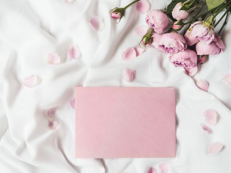 Roses and petals on crumpled white fabric. Natural elegant decoration. Romantic background with copy space on pink envelope. Top view, flat lay.