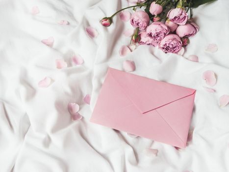 Roses and petals on crumpled white fabric. Natural elegant decoration. Romantic background with copy space on pink envelope. Flat lay.