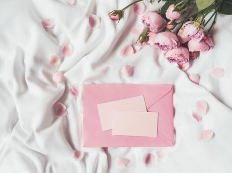 Roses and petals on crumpled white fabric. Natural elegant decoration. Romantic background with copy space on pink envelope and visit cards. Top view, flat lay.