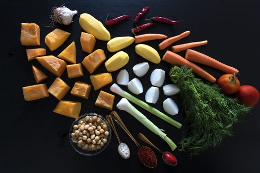 Food on a table. High quality photo. Chopped vegetables on a black background. Pumpkin, potatoes, garlic, chili peppers, carrots, tomatoes, onions. A teaspoon of gold with tomato paste, a teaspoon of gold with Atlantic salt, a teaspoon of gold with paprika, dill, coagulation, a bowl of chickpeas. Ingredients for making couscous.