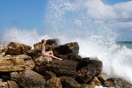 teenage girl sitting on a stone on the seashore under the splashing waves