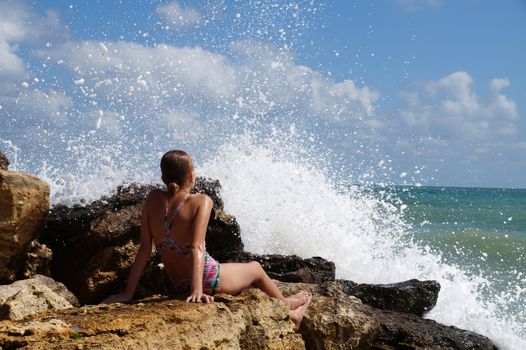 teenage girl sitting on a stone on the seashore under the splashing waves