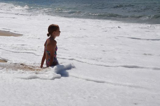 teenage girl sitting in white sea foam and looking at the sea.