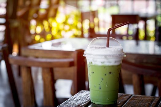 Iced green tea or matcha latte with milk foam in a plastic cup with brown straw on a wooden table in the coffee shop with sun light. Healthy drink and beverage concept.