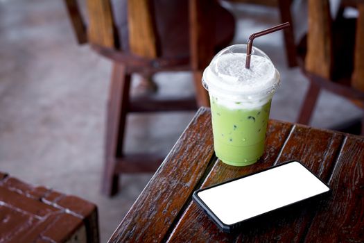 Iced green tea or matcha latte with milk foam in a plastic cup with brown straw and black mobile or smartphone and white scene on a wooden table in the coffee shop. Healthy drink and beverage concept.