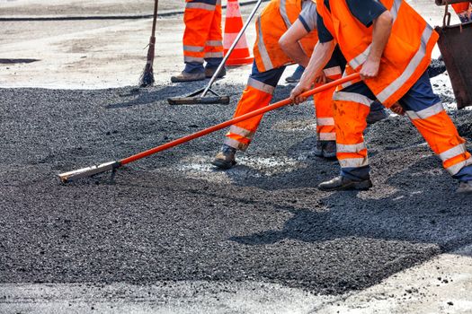 A working group of road workers in orange overalls renews a section of the road with fresh hot asphalt and smooths it out for repair with metal levels.