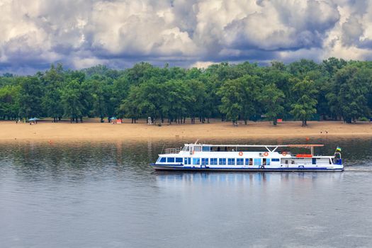 A river tram with a few tourists resting goes along the river against the backdrop of coastal greenery, storm sky and a deserted beach in anticipation of an impending thunderstorm. River passenger transport, concept, copy space.