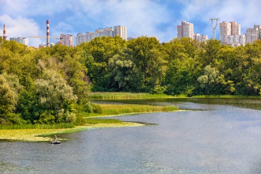 A beautiful bay of the Dnipro with reed-overgrown banks, a fisherman on a boat is catching fish near the shore, and on the horizon is the cityscape and landscape of Kyiv against the background of the blue sky.