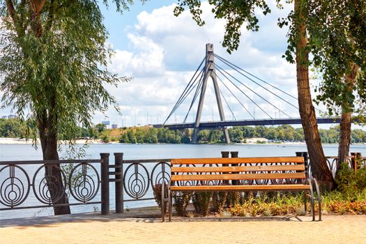 Park wooden bench on the Dnipro embankment against the background of a summer landscape and the Northern bridge over the river in Kyiv.