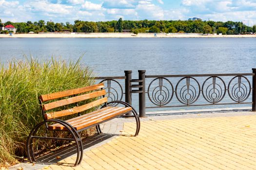 Park wooden bench on the cobbled sidewalk of the Dnipro embankment against the backdrop of a summer landscape and a sandy beach across the river in Kyiv.