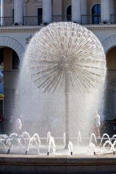 A light refreshing drizzling breeze from the city fountain in the form of a huge dandelion gives the freshness of water coolness to passers-by in the summer heat against the background of the arched-column facade of the Kyiv City Conservatory.