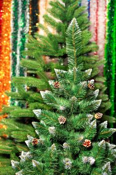Artificial Christmas pine tree with opened cones and white tassels on the branches against a background of multicolored tinsel in blur.