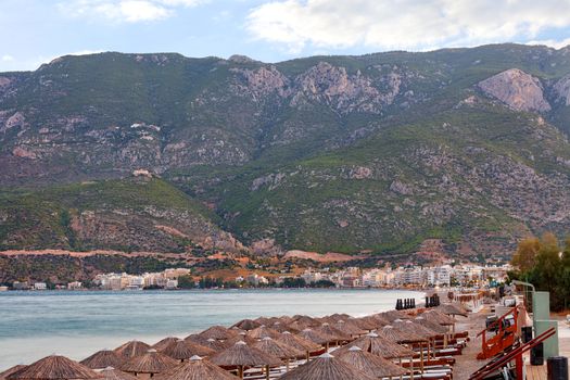 Thatched peaks of beach umbrellas with sun loungers on the deserted shore of the Corinthian Gulf embankment against the backdrop of the morning colors of the awakening city of Loutraki in Greece and mountain ranges in haze and blur.