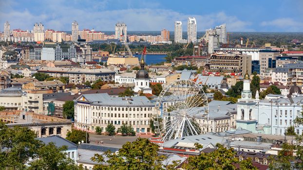 View of the old Podolsky district of the summer city of Kyiv, the old square with a Ferris wheel and a bell tower with a gilded dome, the Dnipro River and many city buildings.