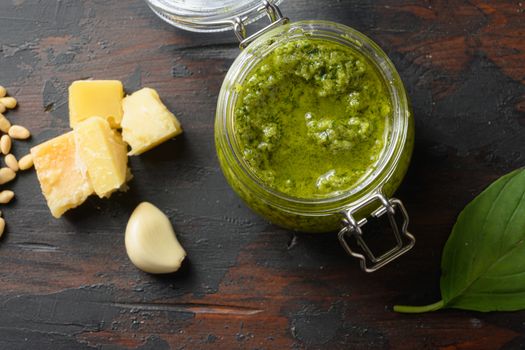 Pesto alla Genovese in glass jar with ingredients parmesan basil pine nuts. on dark wood table planks vintage side view.