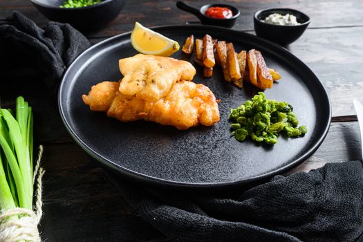 Fish and chips. Traditional british fast food hot dish consisting of fried fish, potato chips, mushy peas and tartare sauce. served on black plate over old dark wood table in pub side view.