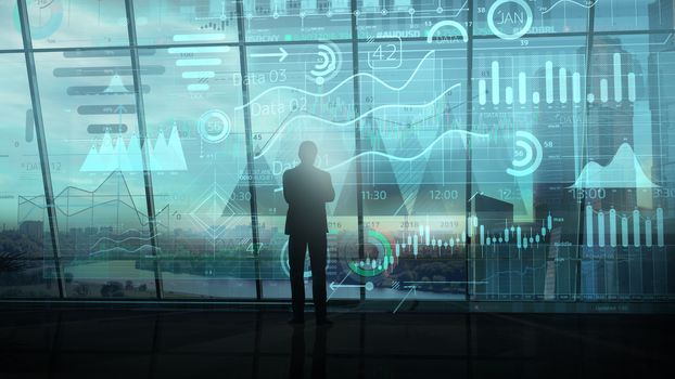 Silhouette of a businessman and a large array of infographics in front of the panoramic window of a dark office.