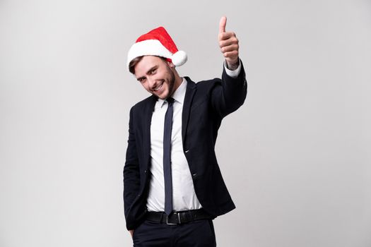 Young handsome caucasian guy in business suit and Santa hats on white background in studio smilie and showing thumbs up two hands. Close up portrait business person with Christmas mood Holiday banner