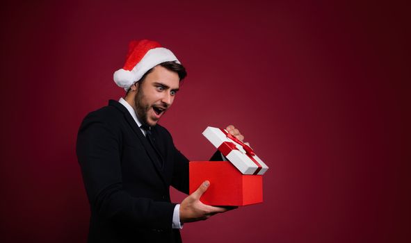 Surprised handsome caucasian guy in business suit and Santa hats stands on white background in studio and smilie opens red gift box in hand. Portrait business person with Christmas mood Holiday banner