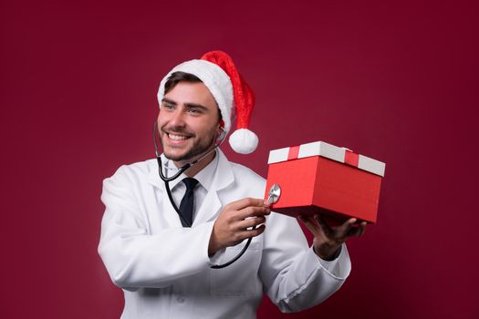 Young handsome doctor in white uniforme and Santa Claus hat standing in studio on red background smile Holding red gift box in hand Portrait medical student Christmas New Year Holiday Medicine concept