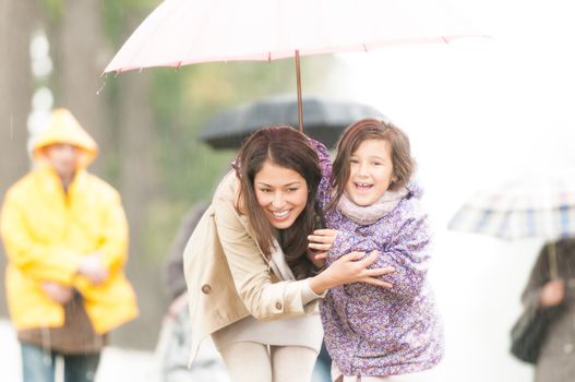 Happy mother and daughter walking in park. Smiling parent and kid hiding under umbrella. Laughing woman and child outdoors. People with umbrellas and person in yellow raincoat in background.