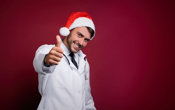 Young handsome doctor in white uniforme and Santa Claus hat standing in studio on red background smile and showing finger in camera Medical student Christmas New Year Holiday Medicine concept