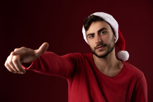 Young handsome caucasian guy in red sweater and Santa hats stands on red background in studio and Shows finger at the camera Close up portrait european guy with Christmas mood Holiday theme. New Year