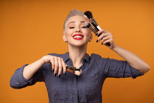 Close up portrait makeup artist. Make up courses. Concept of self visage masterclasess. Woman hold makeup brushes on the hands. Studio shot on the orange color background. Positive people smiling