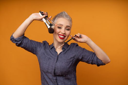 Close up portrait makeup artist. Make up courses. Concept of self visage masterclasess. Woman hold makeup brushes on the hands. Studio shot on the orange color background. Positive people smiling