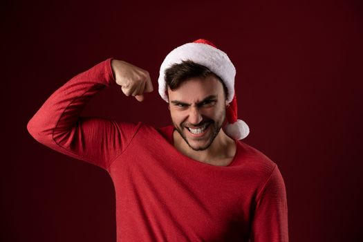 Young handsome caucasian guy in red sweater and Santa hats stands on red background in studio and shows biceps power. Close up portrait european guy with Christmas mood. Holiday theme. New Year.