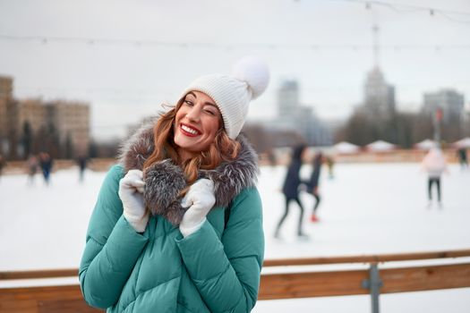 Beautiful lovely middle-aged girl curly hair warm winter jackets white knitted hat glove stands ice rink background Town Square Christmas mood lifestyle Happy holiday woman snowy day Winter leisure