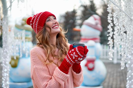 Beautiful lovely middle-aged girl curly hair warm winter sweater red knitted hat glove stands background Town Square Christmas mood lifestyle Happy holiday woman drink hot beverage coffee
