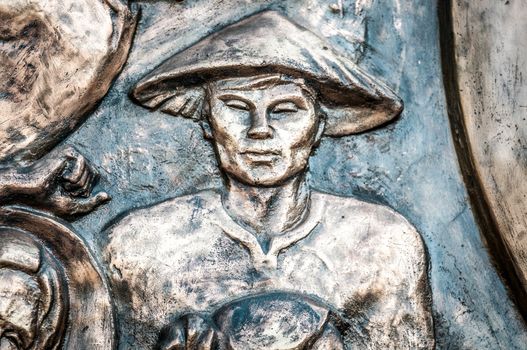 Part of bronze monument with asian man in traditional conical hat. Closeup of metal sculpture with rough dark structure. Beautiful eastern artwork. Vietnam, Asia.