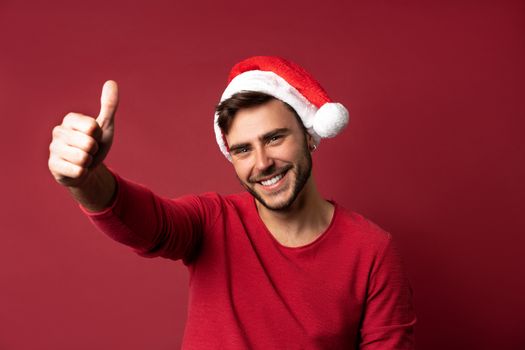 Young handsome caucasian guy in red sweater and Santa hats stands on red background in studio and showing thumbs up. Close up portrait european guy with Christmas mood. Holiday theme. New Year.