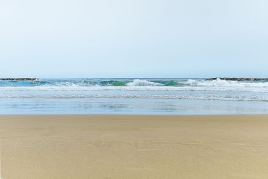 A sandy beach next to the ocean. A calm and quiet beach with deep turquoise clear water. Waves crashing on a breakwater. Soft, golden sea sand without a trace. The reflection of the sky on the water. Clear horizon line. High quality photo