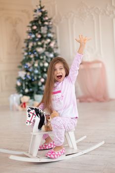A beautiful little girl in pink pajamas rejoices in a wooden rocking horse, a gift from Santa for Christmas.