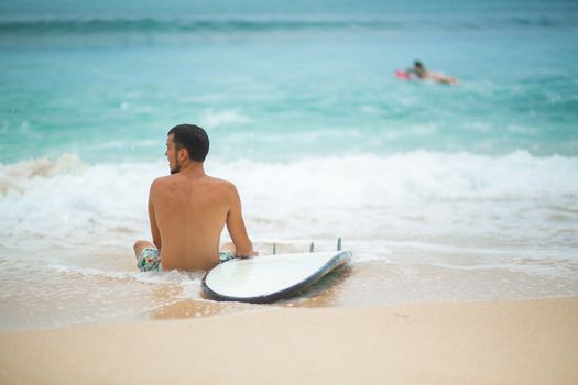 The guy is resting on a sandy tropical beach, after riding a surf. Healthy active lifestyle in summer vocation.